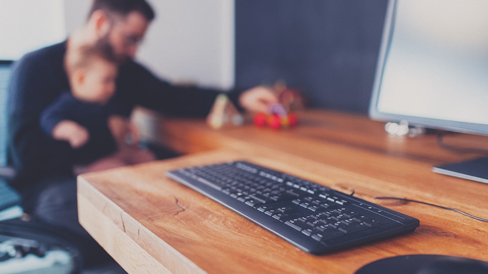 Arbeitsplatz mit Tastatur, während eine Person mit Kind im Hintergrund spielt.